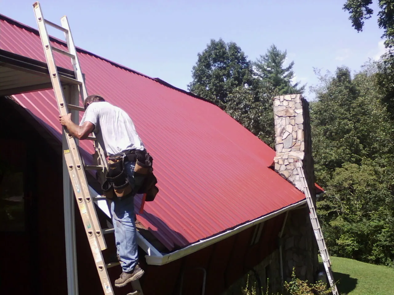 Roofer on a ladder beside a red metal roof and stone chimney — Soffit & Fascia Repair by Roofing Brothers in Burlison